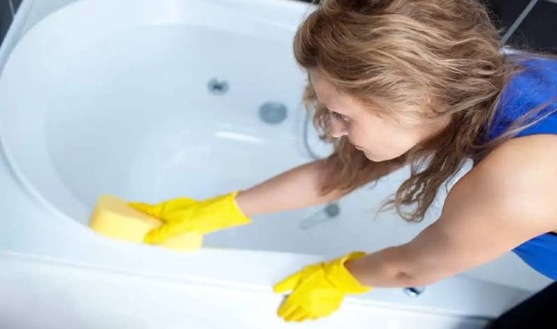 Women in blue dress and yellow gloves scrubbing bathtub