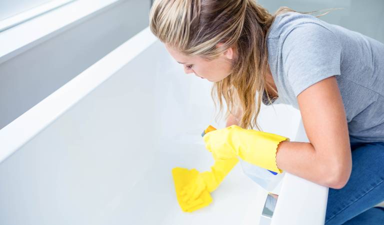 Woman in yellow gloves scrubbing bathtub and holding bottle in her hand