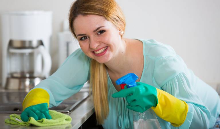 Woman in blue top and green-yellow gloves holding a bottle and cloth inside a kitchen