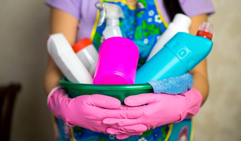 Woman in purple top holding a green basket filled with tools, bottles, supplies etc.