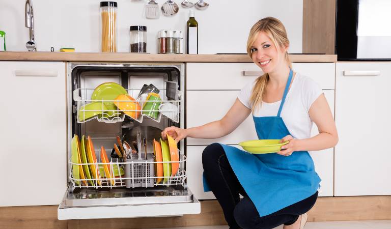 Woman in white blue dress holding a plate in her hand beside dishwasher