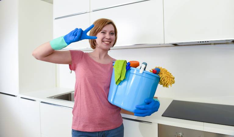 Woman in pink top and blue gloves holding a blue basket filled with tools, supplies etc.