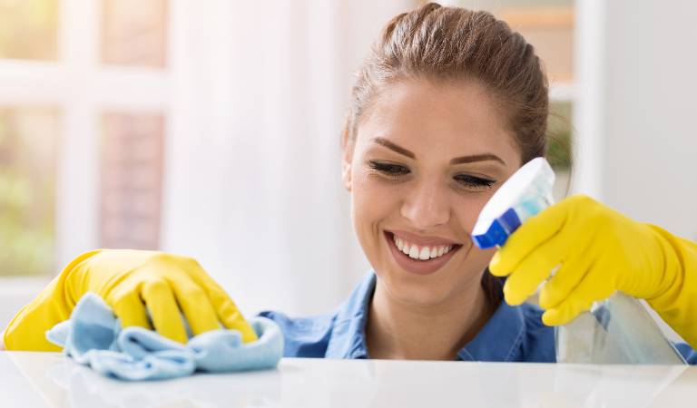 A smiling woman in yellow gloves holding a spray bottle in her hand and cleaning a table with a cloth.