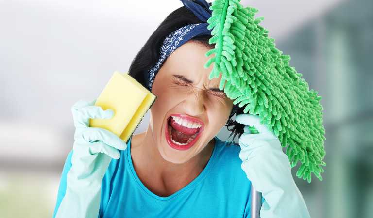 Irritate woman in blue top holding a yellow scrubber and green cleaning brush shouting.