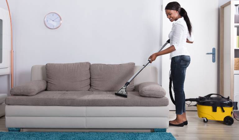Woman in white top and blue jeans cleaning sofa with vacuum cleaner.