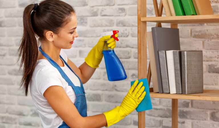 Beautiful woman in white-blue uniform and yellow glove holding a blue bottle in her hand cleaning shelves in a room