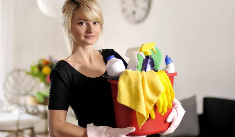 Woman in black dress holding a red basket filled with tools, bottles, cloth.