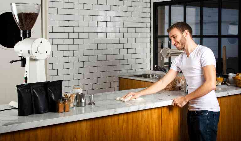 Man in white top and black pant cleaning countertop with a cloth inside his kitchen.
