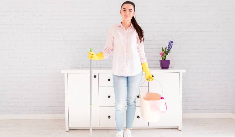 Woman holding a mop and a basket in her hand standing in a room.