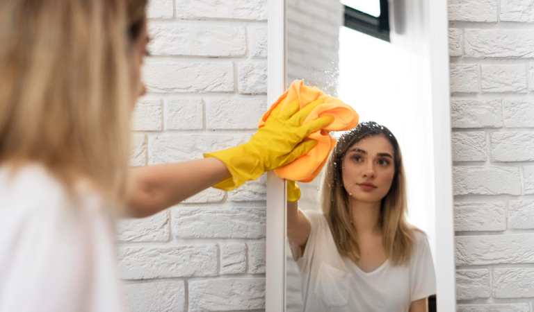 Woman in white top and yellow glove removing soap scum from mirror