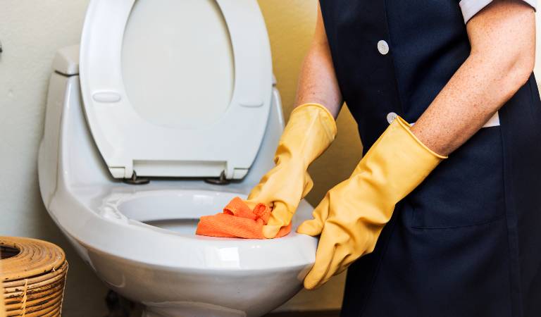 Woman in black uniform and orange gloves cleaning toilet bowl using orange cloth.