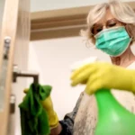 Old woman in green mask holding a green bottle in her hand and cleaning door with a cloth.