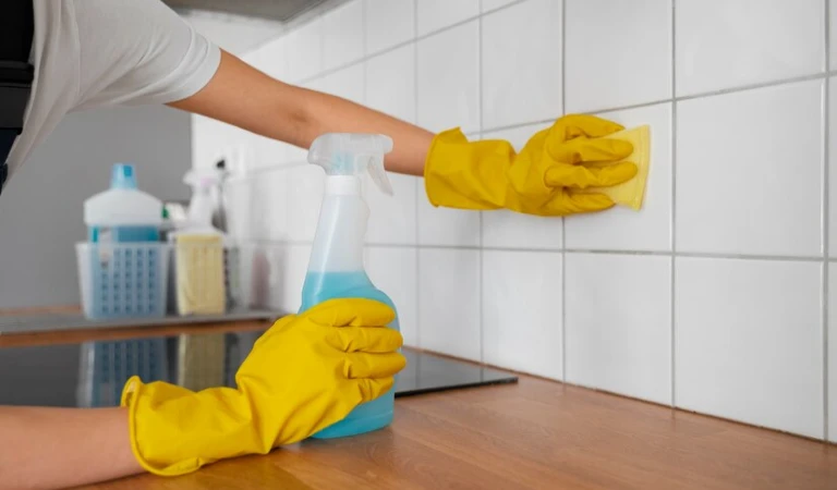 A lady in yellow gloves holding a bottle in her hand cleaning tiles.