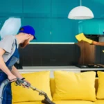 Young man with vacuum cleaner washing with vapor yellow sofa and beautiful woman wipes cuisine furniture