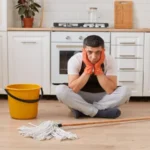 Portrait of sad upset man wearing casual clothing apron and orange rubber gloves sitting on floor in kitchen with crossed legs