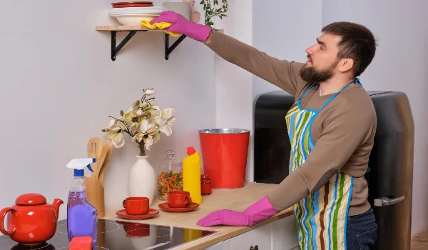 Young bearded man in the kitchen wearing apron and pink gloves cleans the shelf